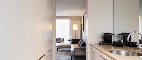 A modern hotel room with a kitchenette, a dining area with a sofa and chairs, and a large window with curtains.