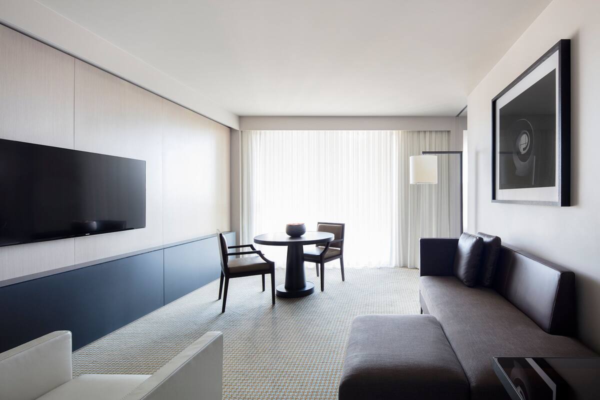 A modern hotel room with a flat-screen TV mounted on the wall, a sofa, a dining table with chairs, and a large window with curtains.