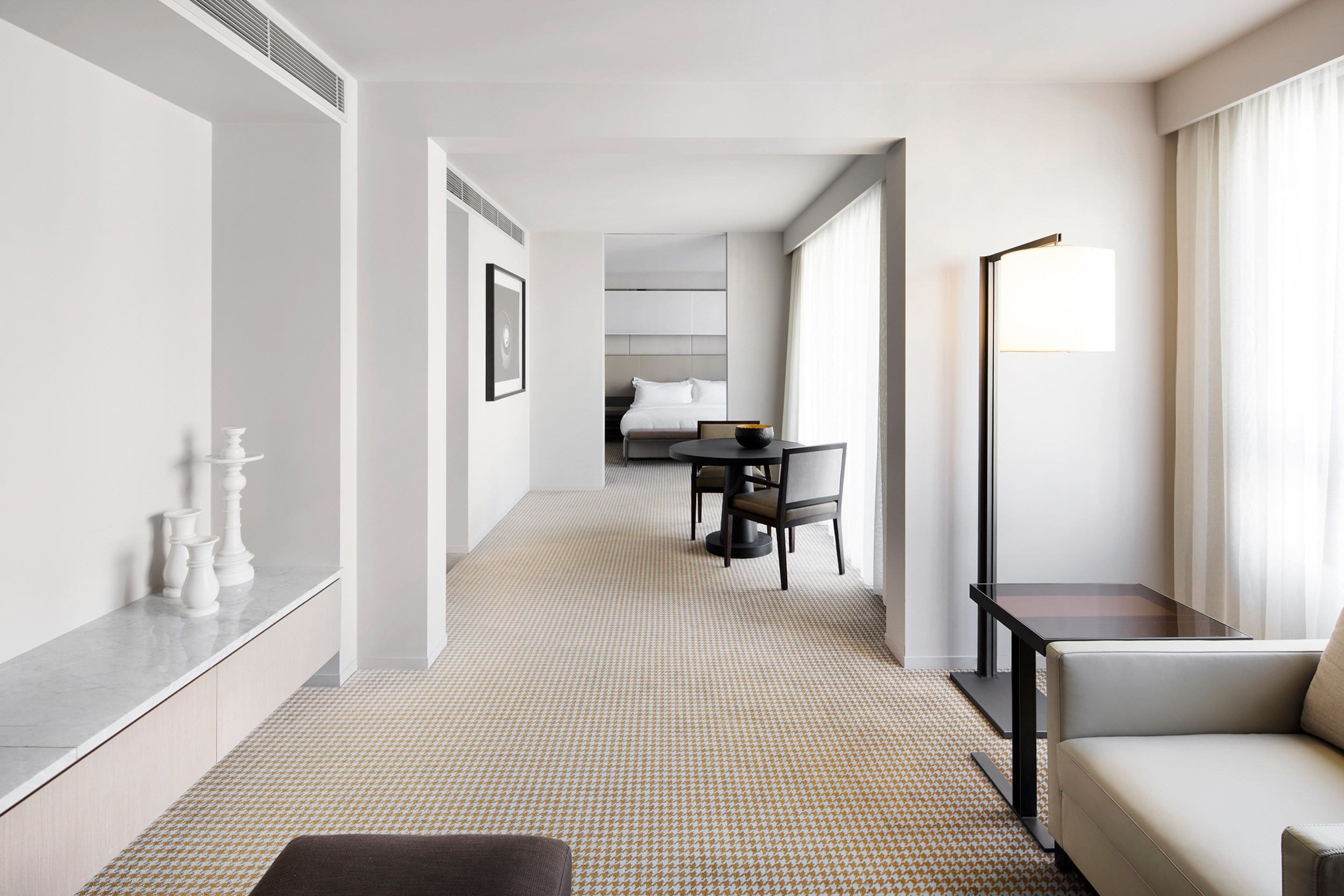 A hotel room with a modern design, featuring a sofa, a dining table with chairs, a lamp, and a bed in the background.