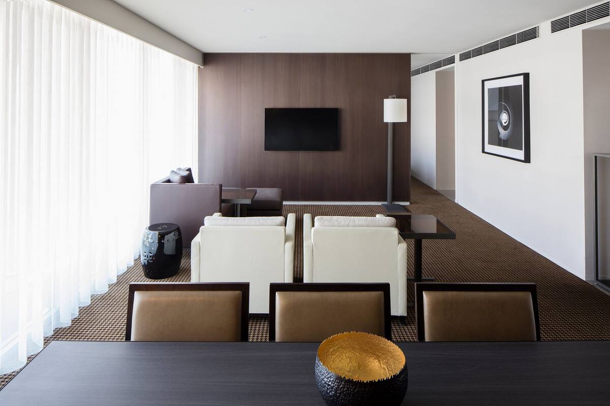A hotel room with a dark wooden accent wall, a flat-screen TV, a sofa, a dining table with chairs, and a large window with sheer curtains.