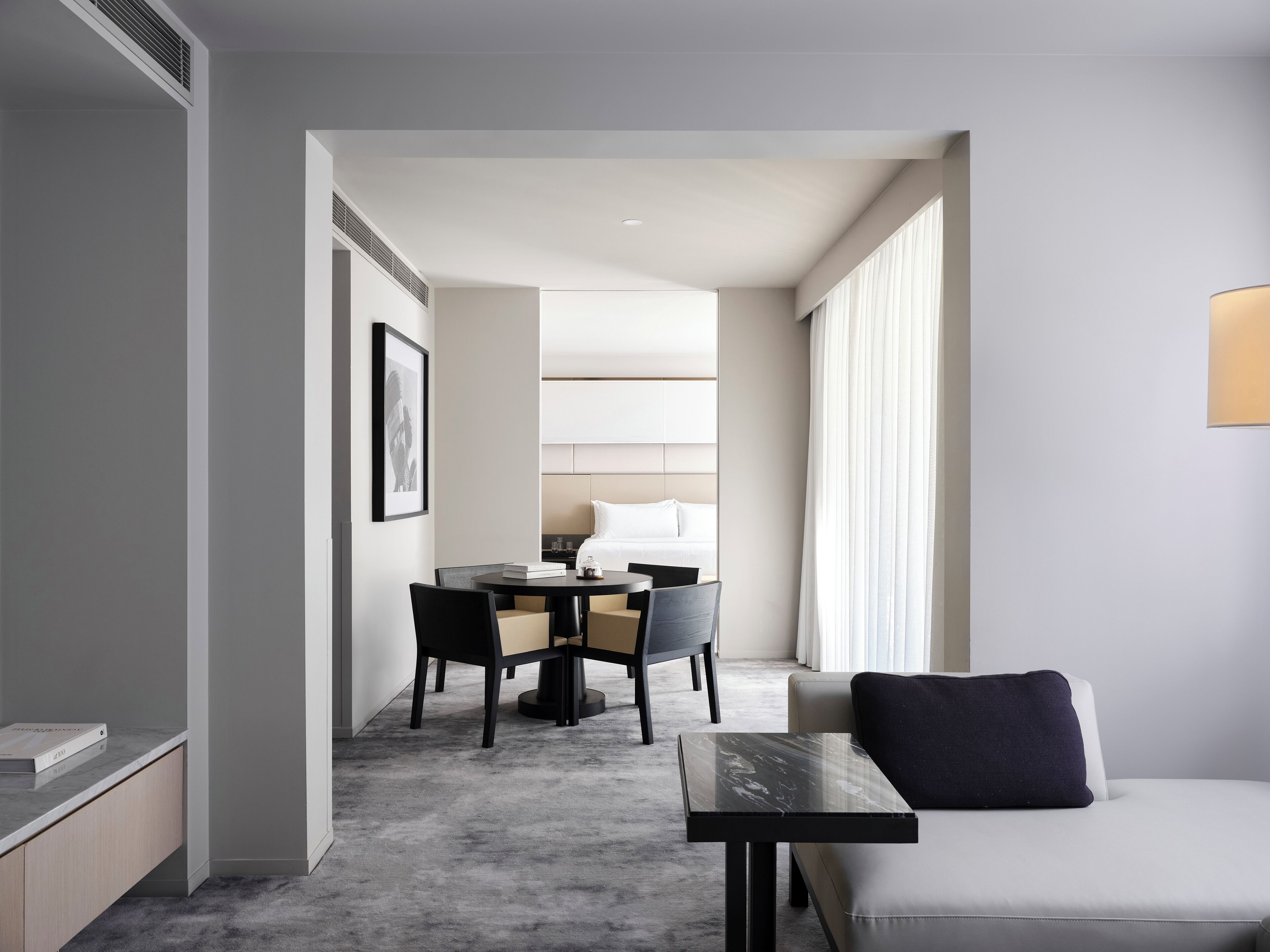 A modern hotel room with a minimalist design, featuring a sofa, a small table, a dining area with chairs, and a large window providing natural light.