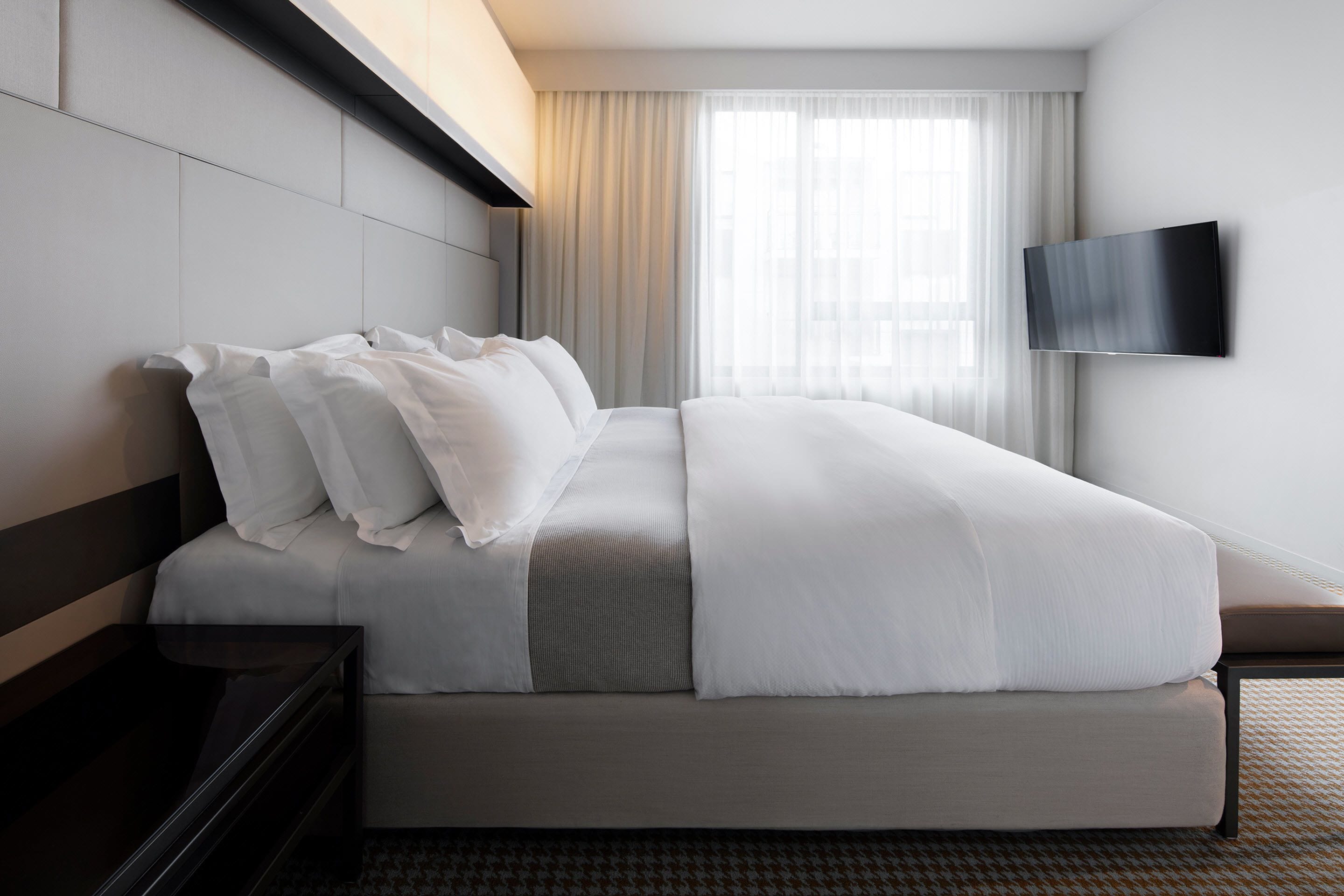 A modern hotel room with a large bed, a flat-screen TV mounted on the wall, and a window with sheer curtains.