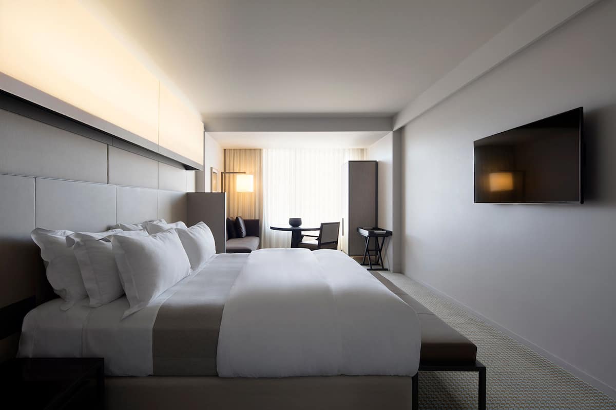 A modern hotel room with a large bed, a flat-screen TV mounted on the wall, a small table with a chair, and a window with curtains.