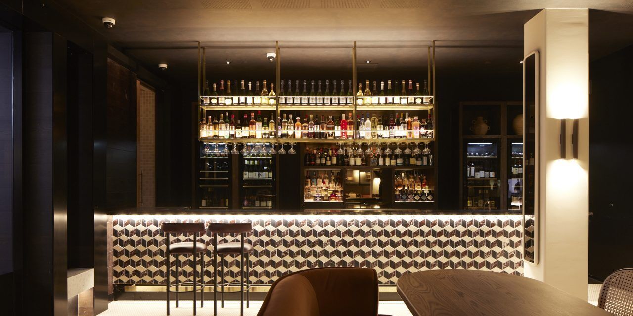 Una zona de bar con una estantería alta de botellas, un área de asientos con un revestimiento de pared estampado y una barra.