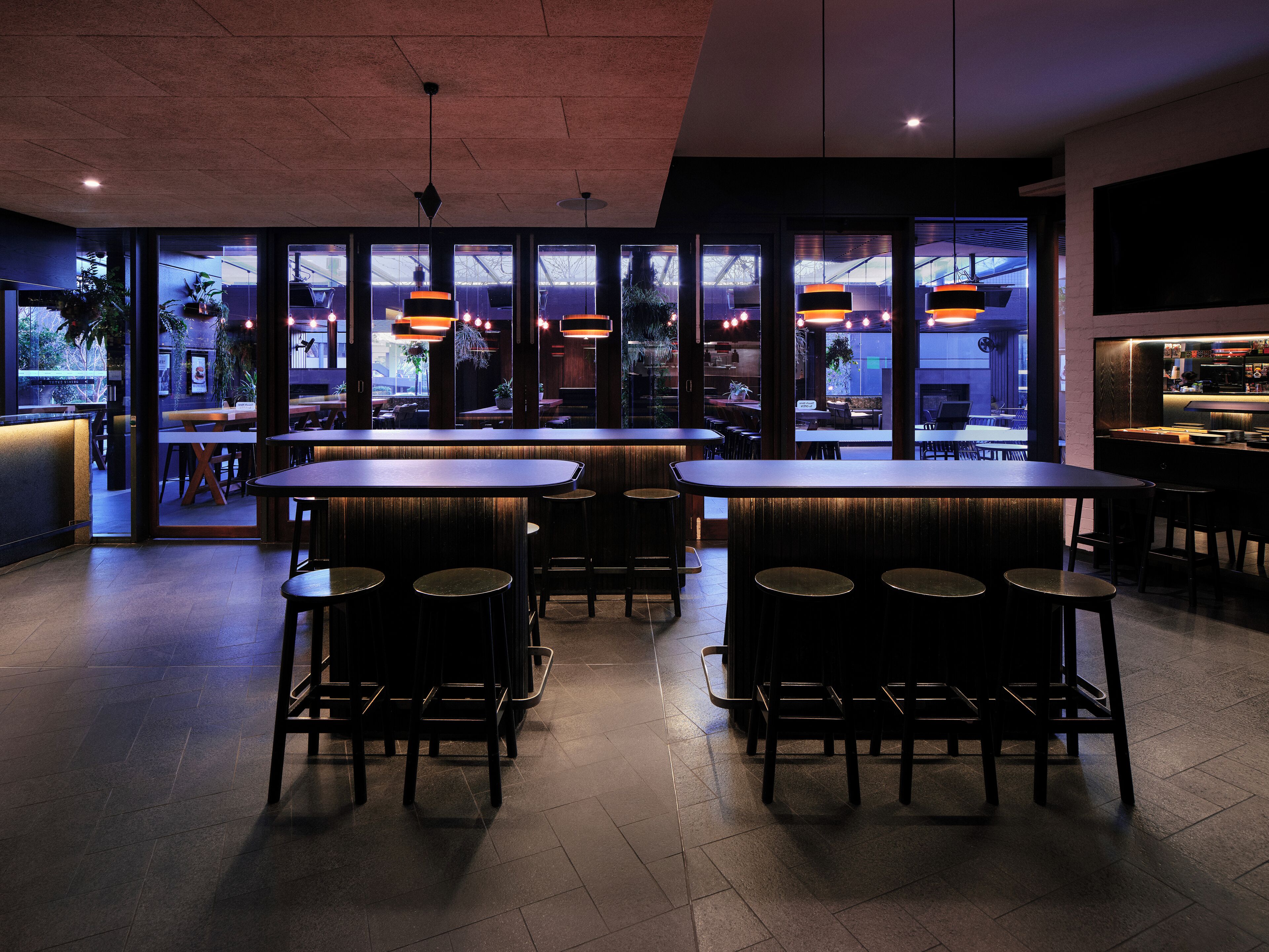 Un área de bar moderna con mesas y taburetes altos, ventanas amplias e iluminación colgante.