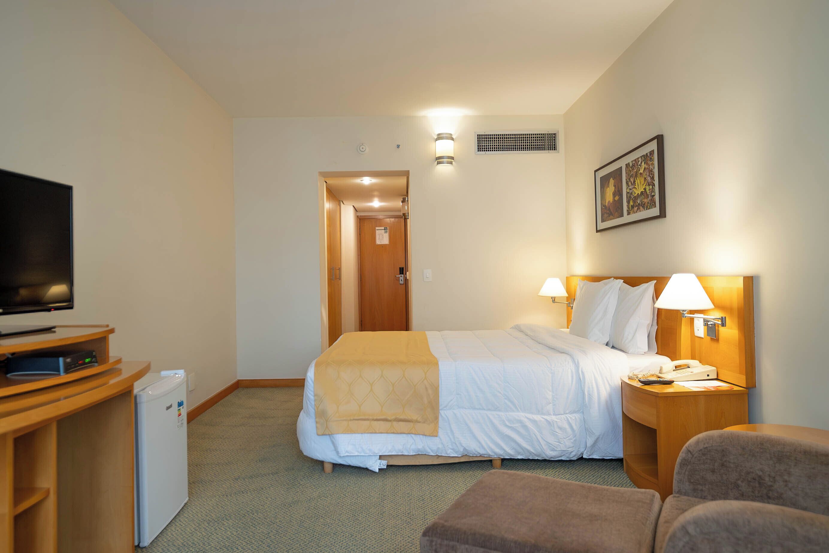Une chambre d’hôtel comprenant un lit, un bureau avec une télévision, un réfrigérateur, une lampe de chevet et un tableau au mur.