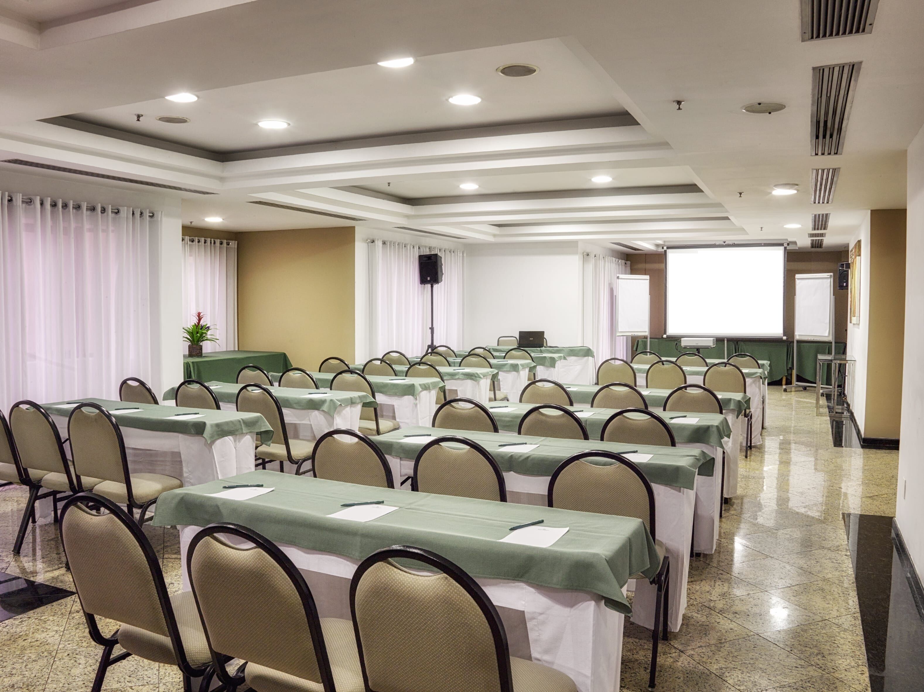 A conference room with rows of tables and chairs, a projector screen, and a speaker on a stand.