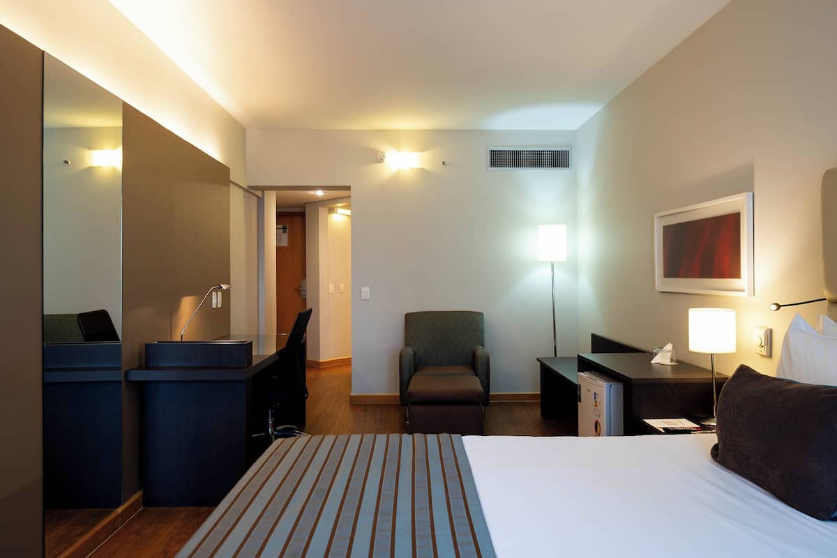 Une chambre d’hôtel comprenant un lit, un bureau, une chaise, un miroir et une table de chevet avec une lampe.