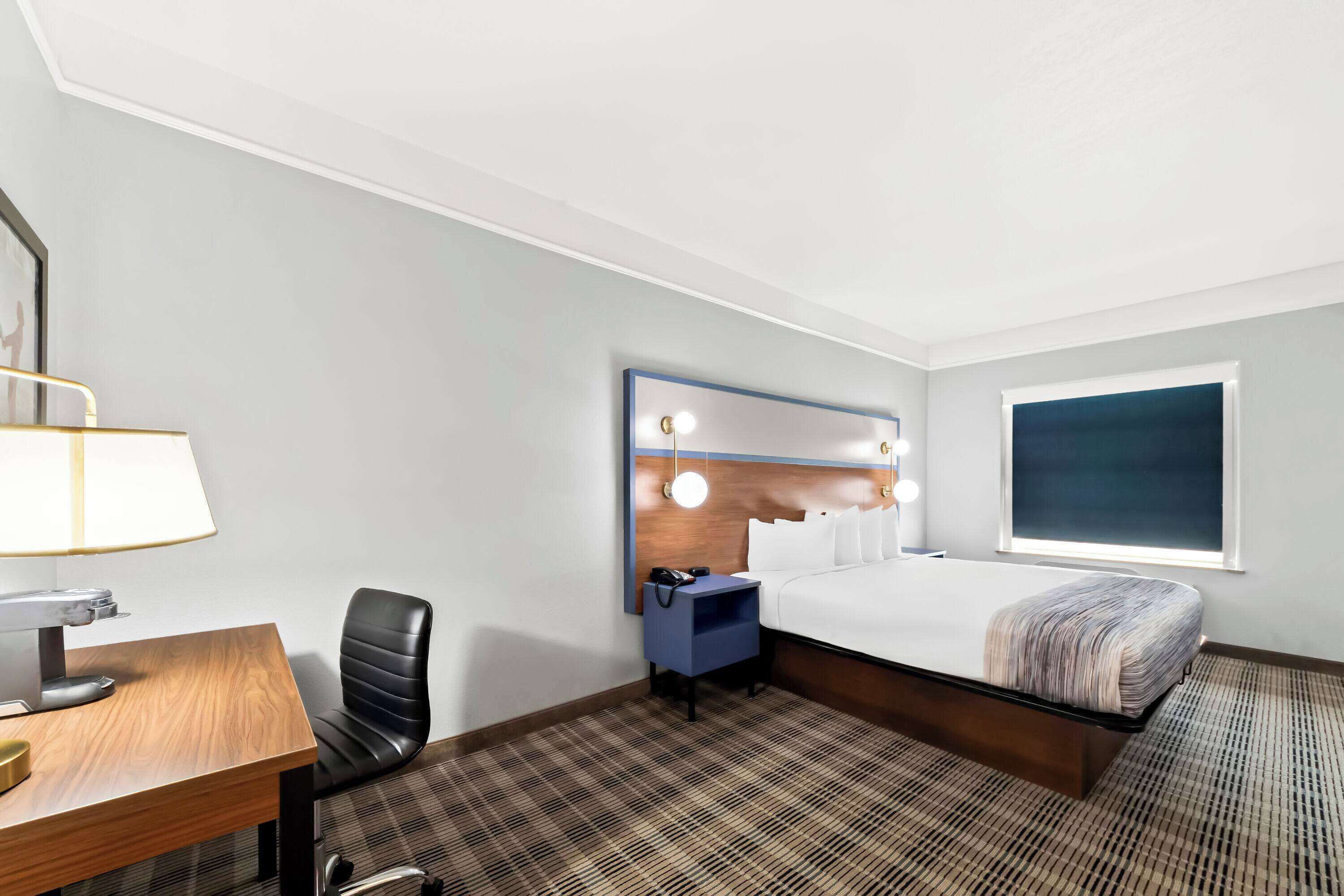 A hotel room with a bed, a desk, a chair, a nightstand, and a mirror with lights.