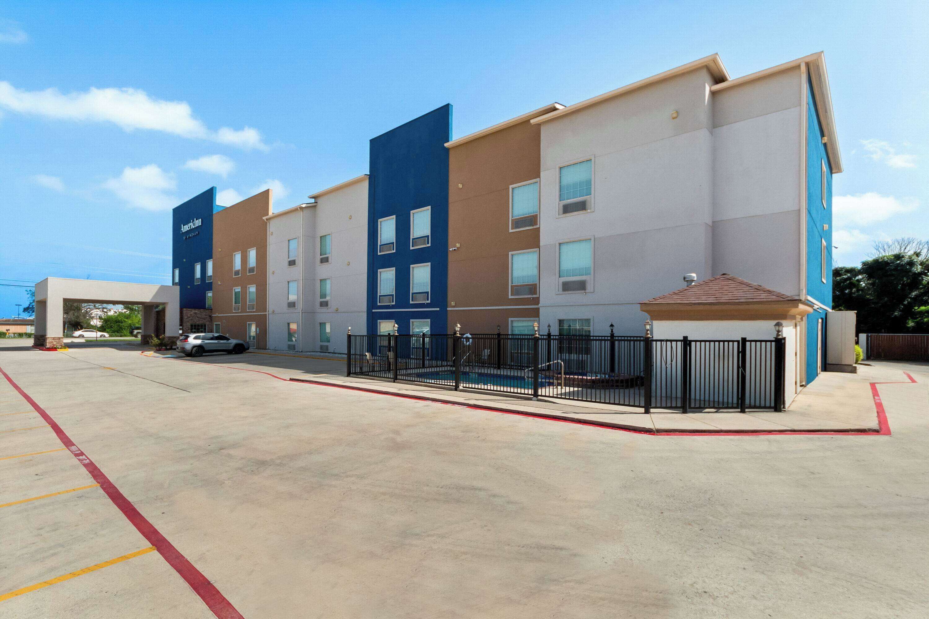 A multi-story hotel with a modern design, featuring a mix of blue and beige facades, a gated entrance, and a parking area with marked spaces.