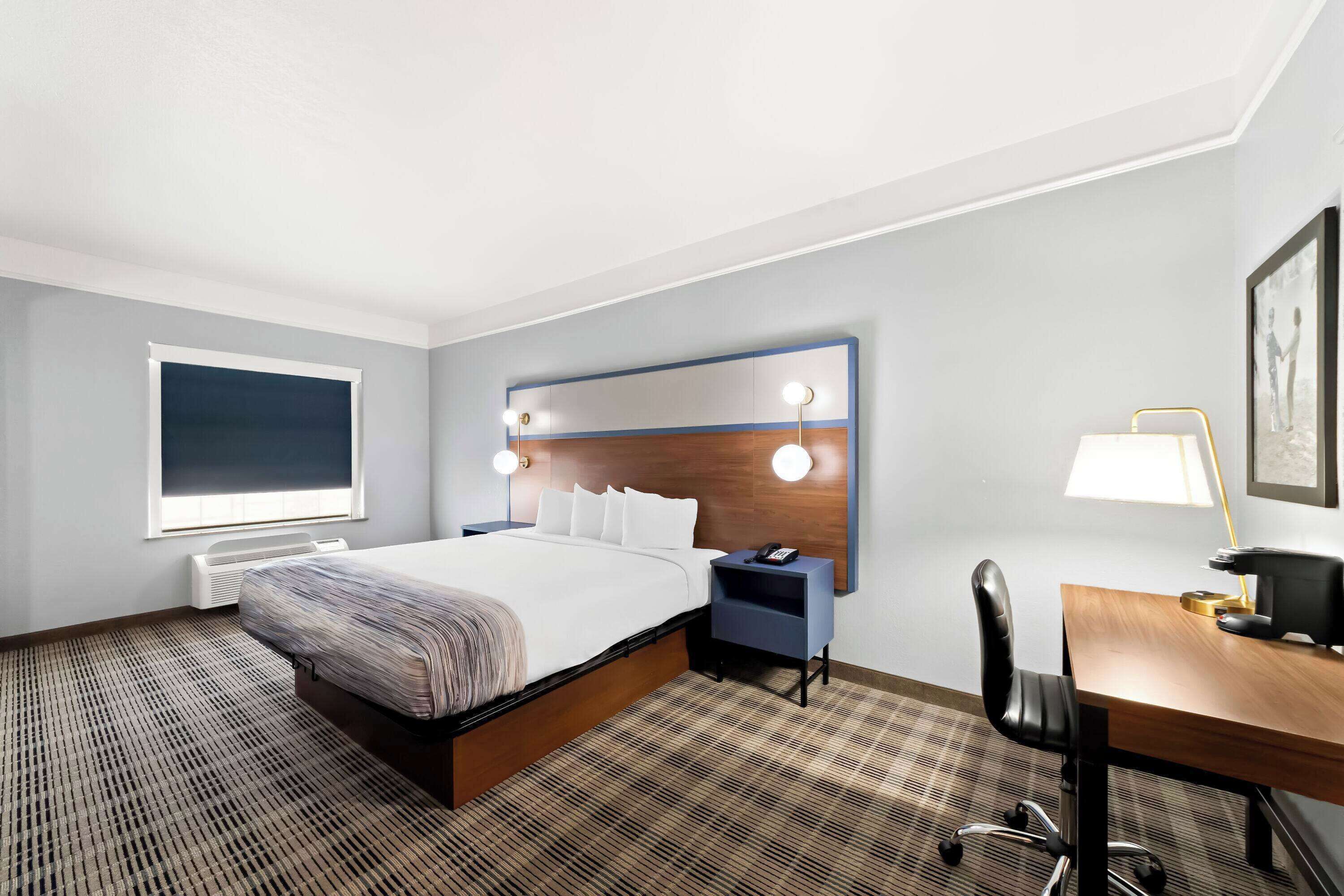 Una habitación de hotel con una cama, mesitas de noche, un escritorio, una silla, una ventana y una lámpara fijada a la pared.
