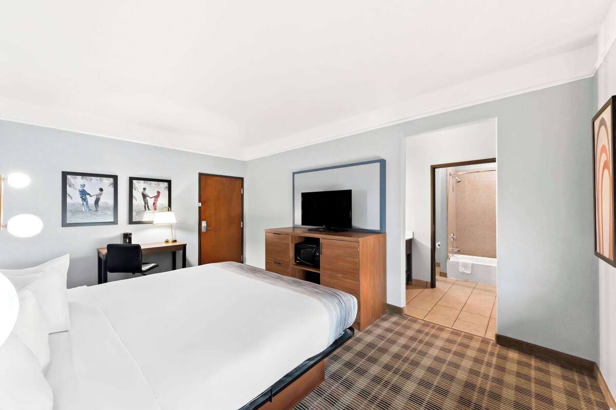 A hotel room with a large bed, a desk with a lamp, a television on a wooden cabinet, and a bathroom visible through an open door.