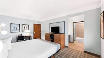 A hotel room with a large bed, a desk with a lamp, a television on a wooden cabinet, and a bathroom visible through an open door.