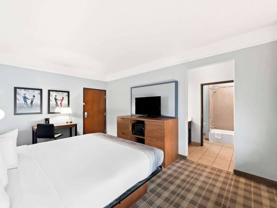 A hotel room with a large bed, a desk with a lamp, a television on a wooden cabinet, and a bathroom visible through an open door.