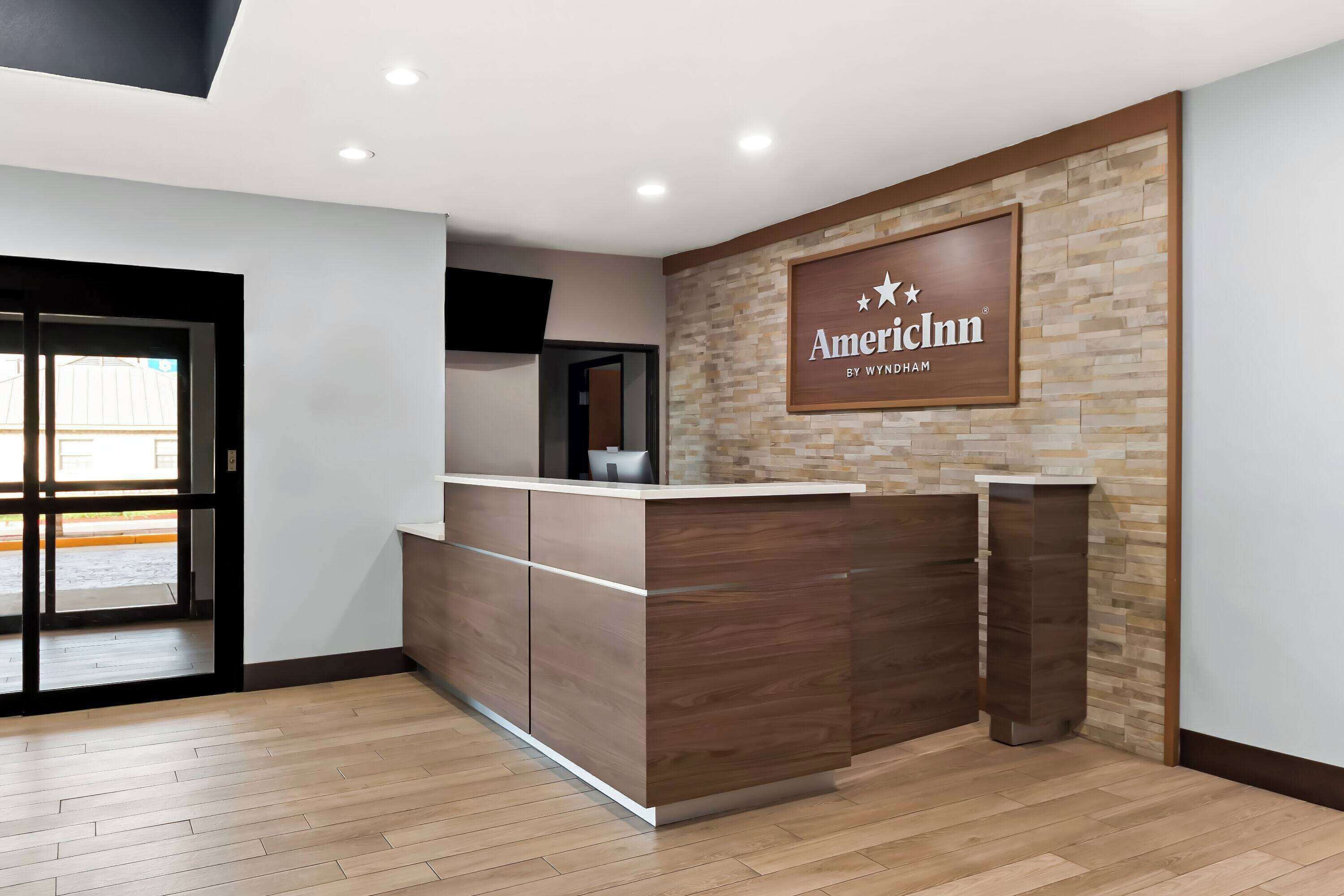 Un mostrador de recepción con un letrero que dice "AmericInn by Wyndham" en un vestíbulo moderno con piso de madera y una pared de acento de piedra.