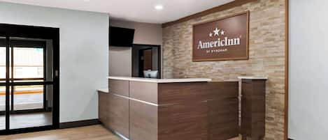 A reception desk with a sign reading "AmericInn by Wyndham" in a modern lobby with wooden flooring and a stone accent wall.