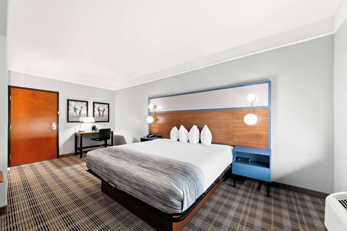 A hotel room with a large bed, a desk, a chair, a desk lamp, a nightstand, and a wall-mounted shelf with decorative items.