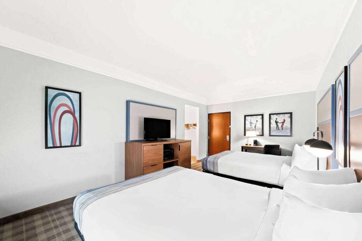 A hotel room with two beds, a TV, a desk with a lamp, and artwork on the walls.