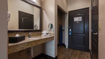 A hotel bathroom with a granite countertop sink, a large mirror, a towel dispenser, and a dark wooden door.