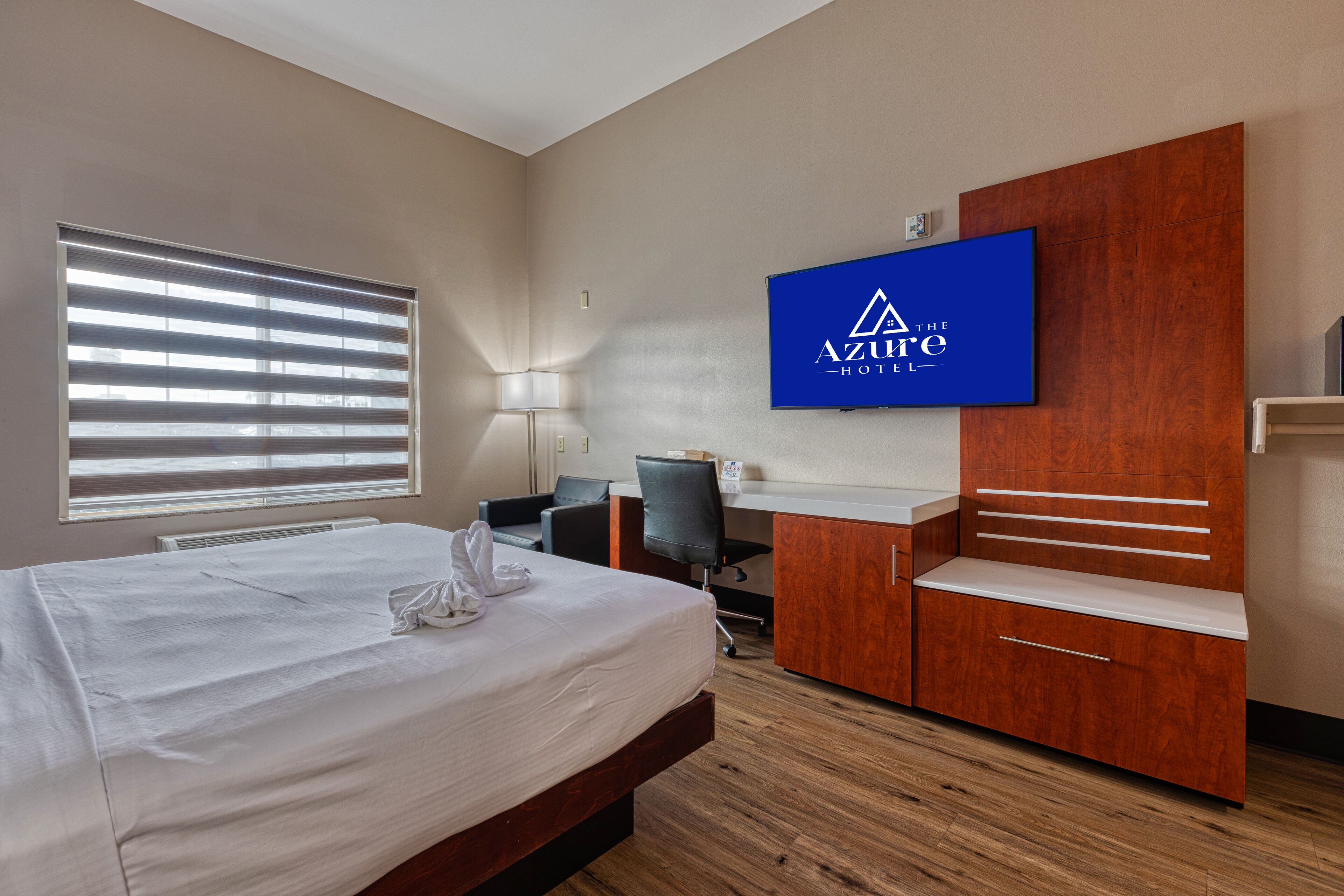 A hotel room with a bed, a desk, a chair, a TV mounted on the wall, and a window with blinds.