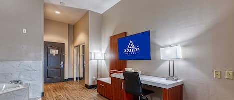 A hotel room with a wooden floor, a desk with a chair, a large mirror, and a television mounted on the wall.