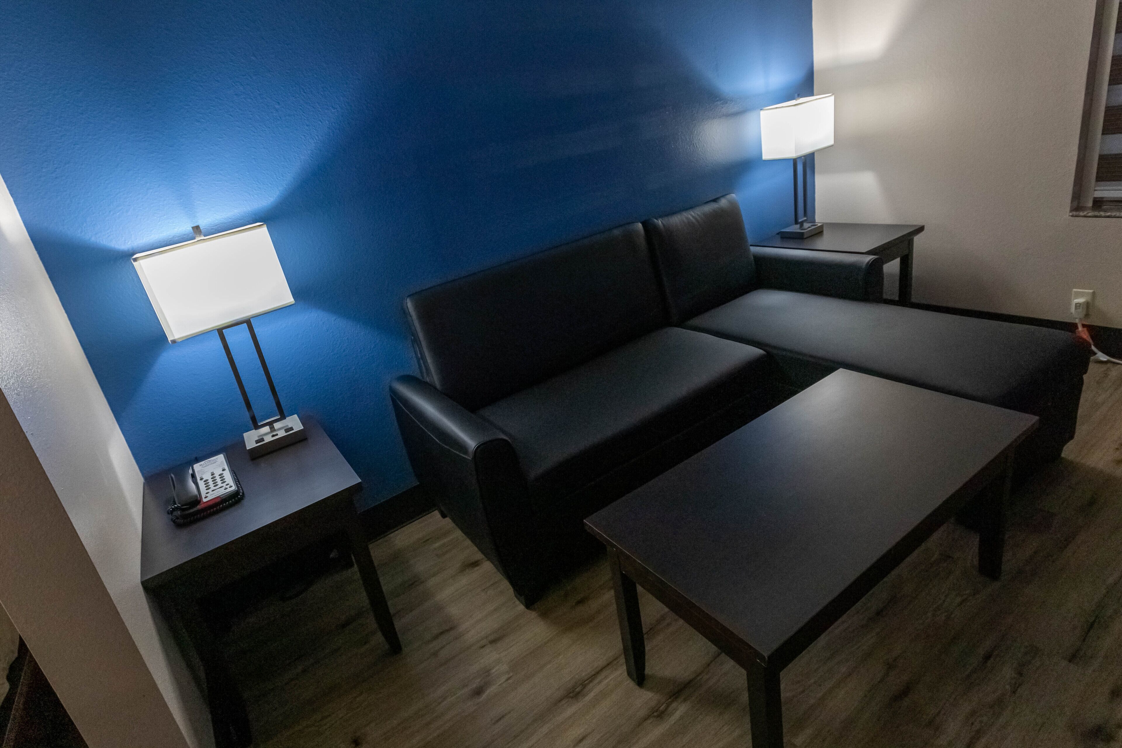 Quarto com sofá preto, uma mesinha, luminária e telefone em uma mesinha lateral.