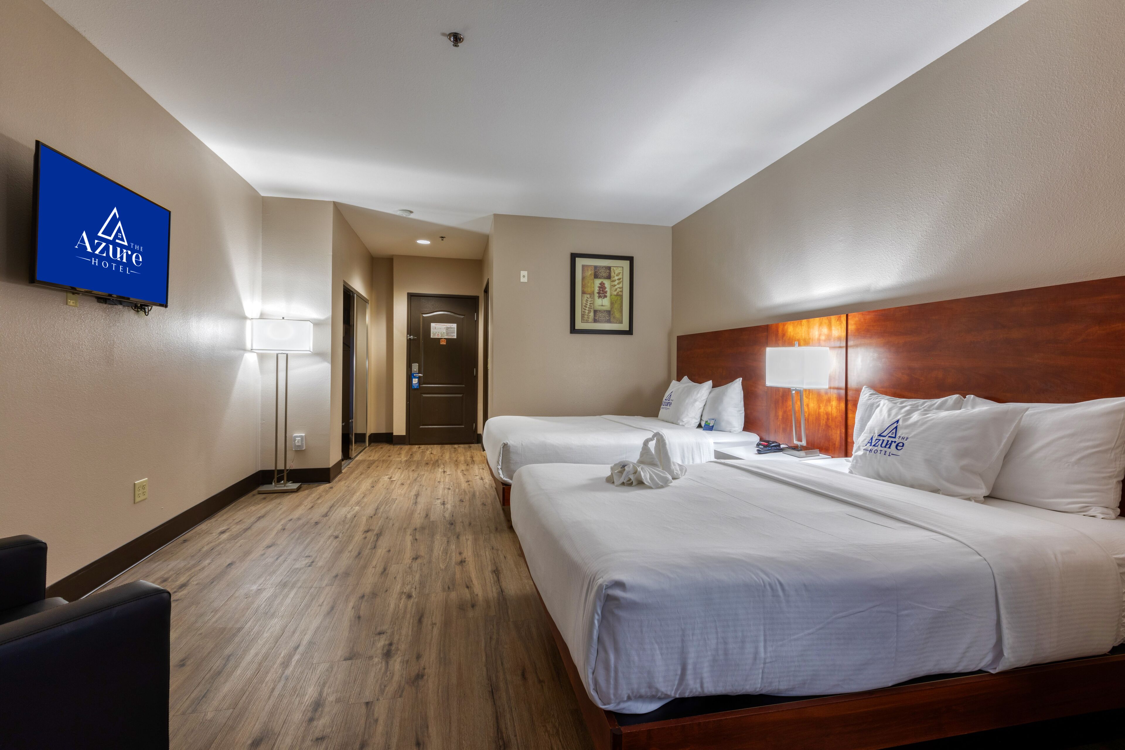 A hotel room with two beds, a flat-screen TV, a wooden floor, and a framed picture on the wall.