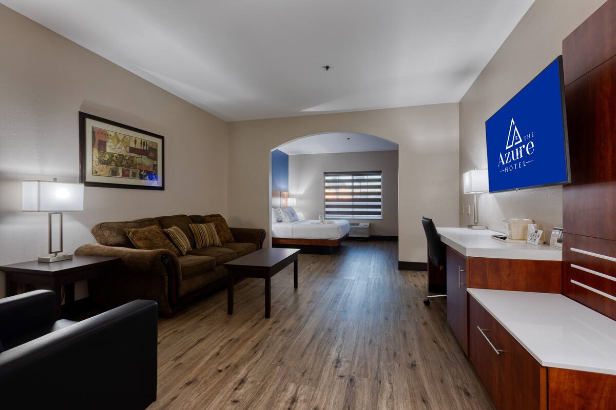 Une chambre d’hôtel avec un canapé marron, une petite table, un bureau avec une lampe, un lit près d’une fenêtre et un coin cuisine.