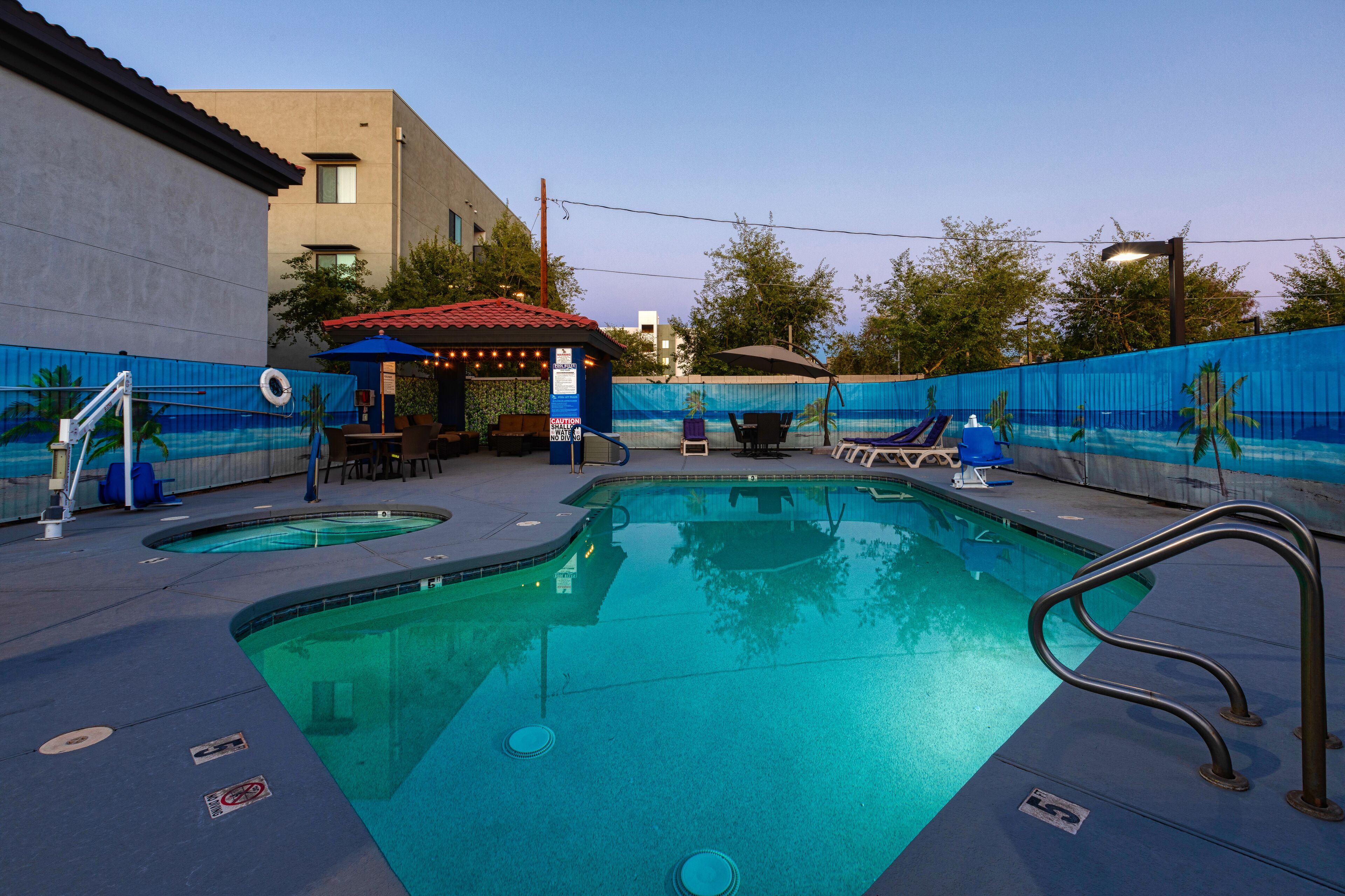 A swimming pool with lounge chairs and umbrellas, surrounded by a blue fence and a building with a red roof.