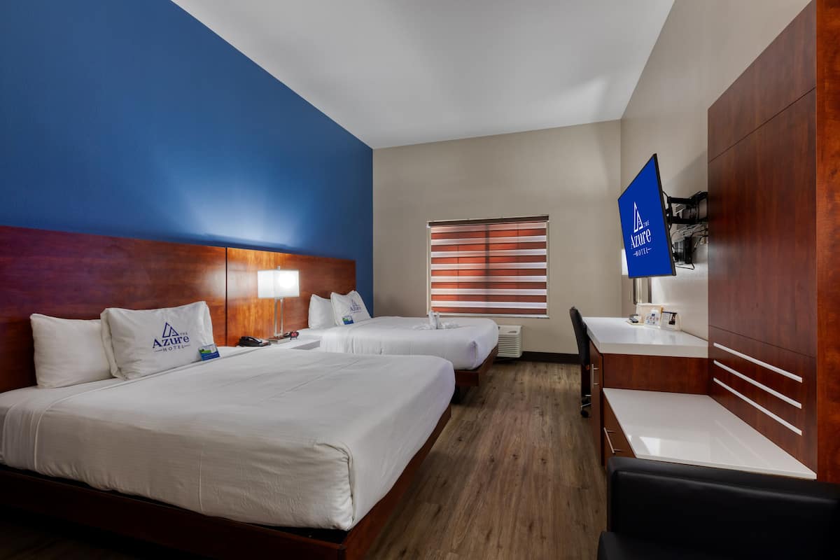 Une chambre d’hôtel avec deux lits, un bureau, une chaise, une fenêtre avec des stores et un panneau bleu sur le mur.