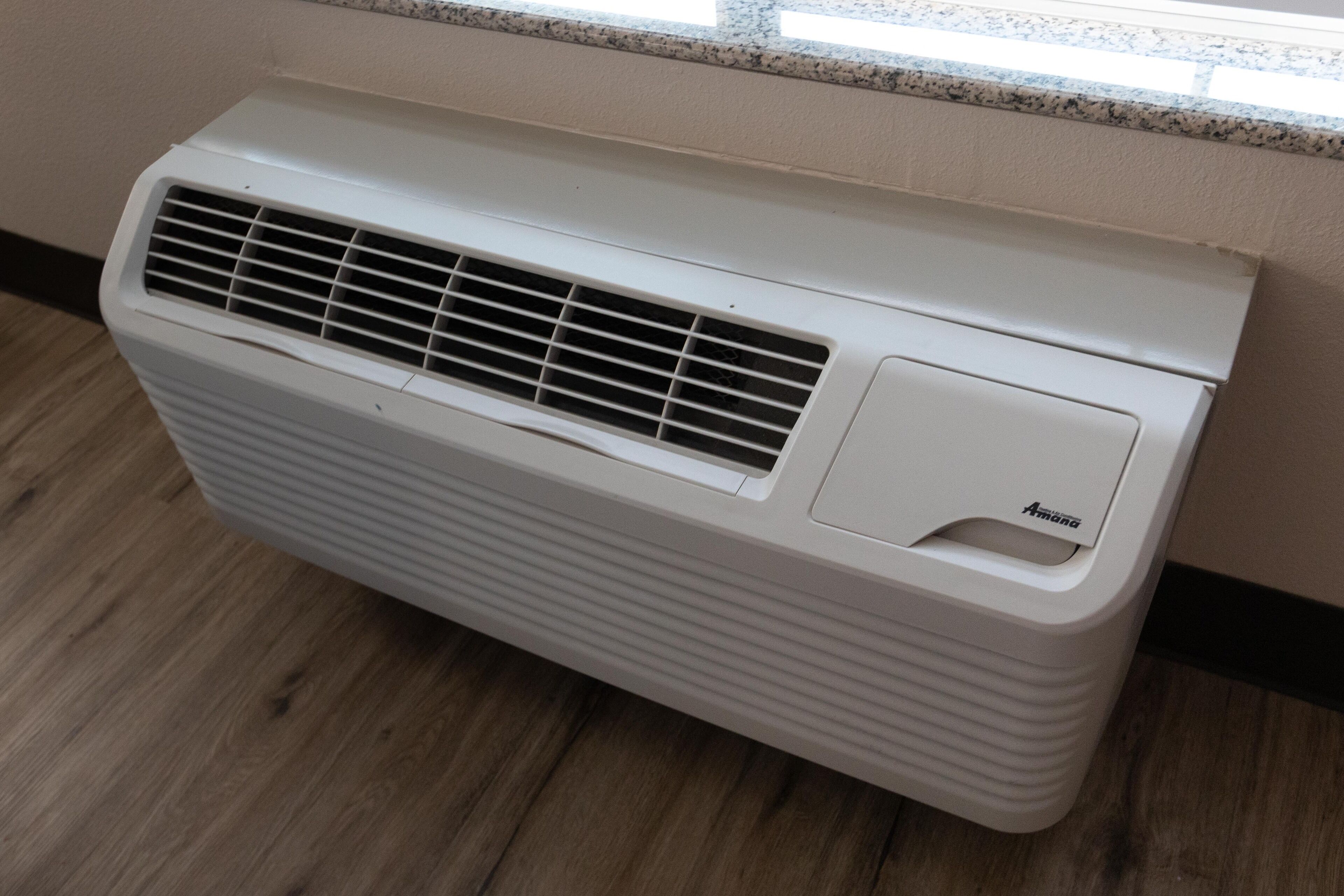 A white air conditioner unit with a vent and a brand label, placed on a wooden floor against a wall.