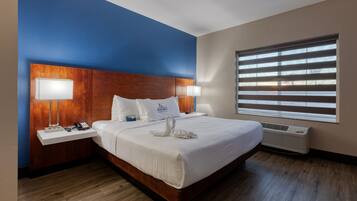 A hotel room with a large bed, two bedside tables with lamps, a wooden headboard, and a large window with blinds.