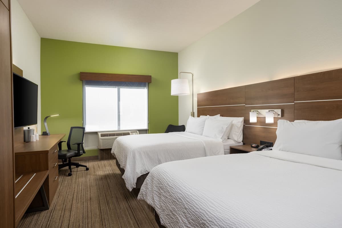 Ein Hotelzimmer mit zwei Betten, einem Schreibtisch mit Fernseher, einem Sessel und einem Fenster mit Jalousien.