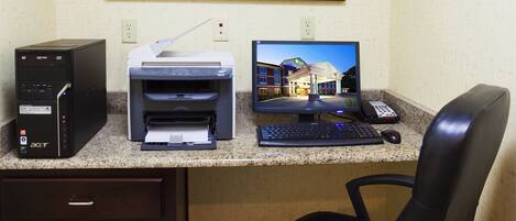 A hotel room with a desk, printer, computer, and a framed floral artwork on the wall.