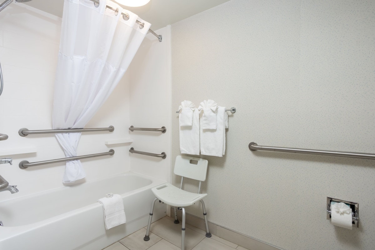 Ein Badezimmer mit einer Badewanne, einer Duschvorhang, einem weißen Stuhl und Handläufen.