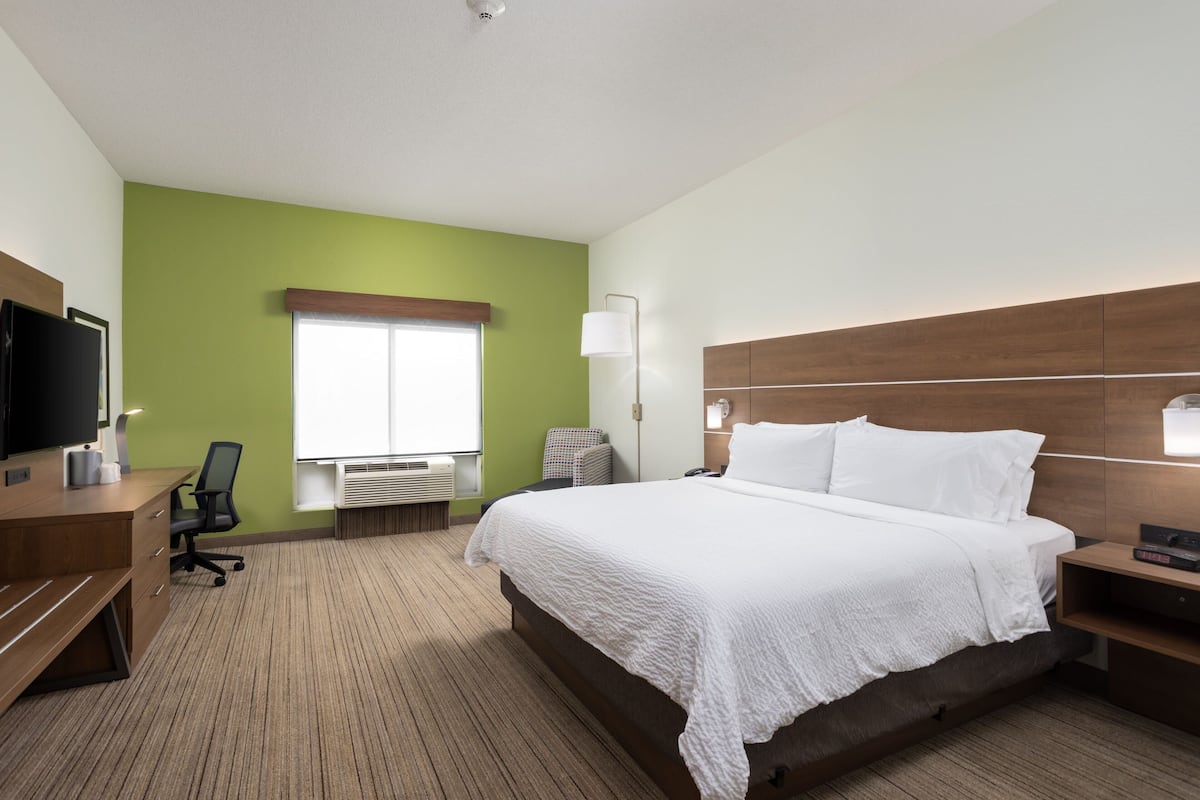 Ein Hotelzimmer mit einem großen Bett, einem Schreibtisch, einem Flachbildfernseher, einem Sessel, einer Lampe und einem Fenster mit Jalousien.
