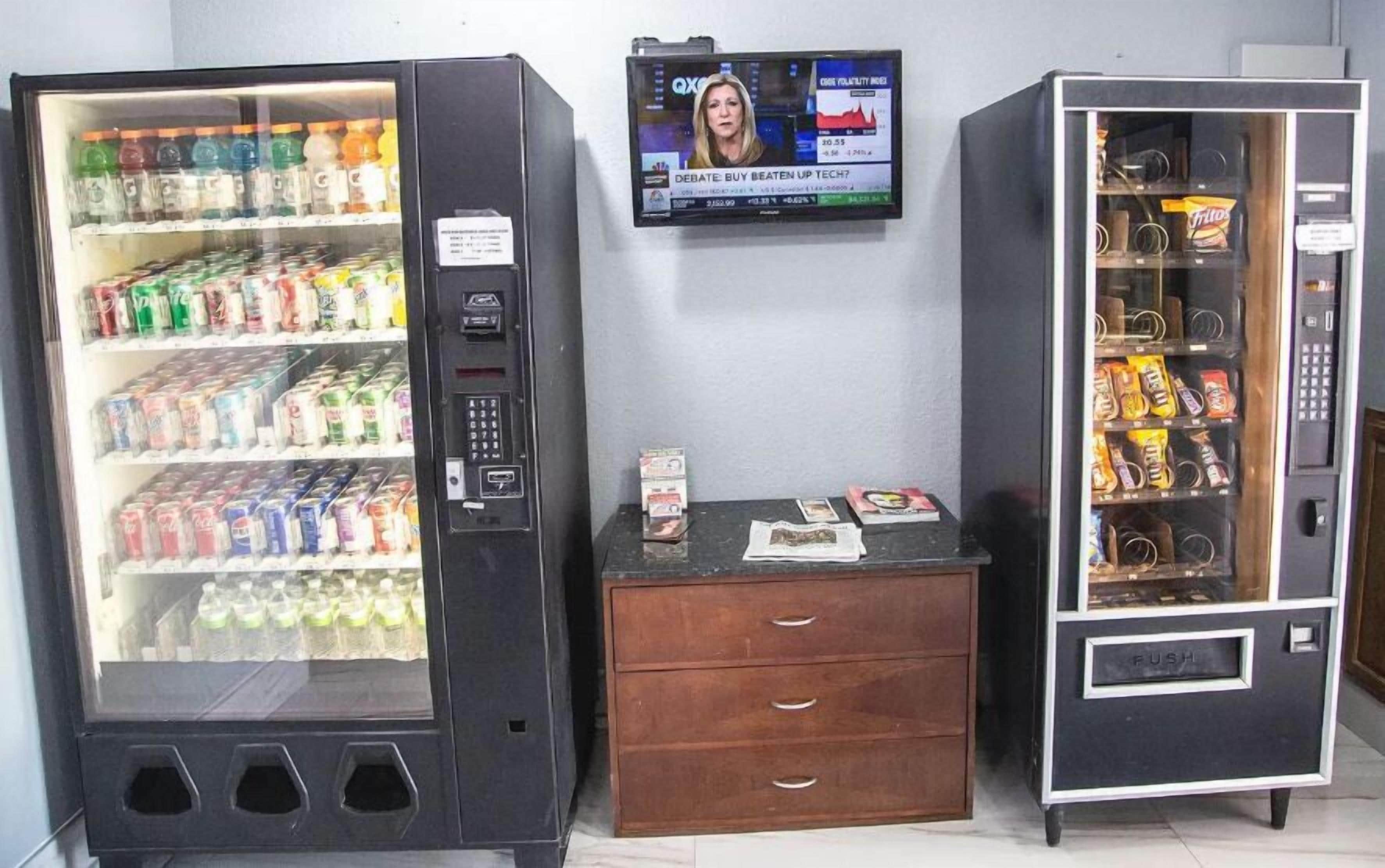 Una máquina expendedora con una pantalla que muestra un programa de noticias, una pequeña mesa de madera con un periódico, y otra máquina expendedora de aperitivos.