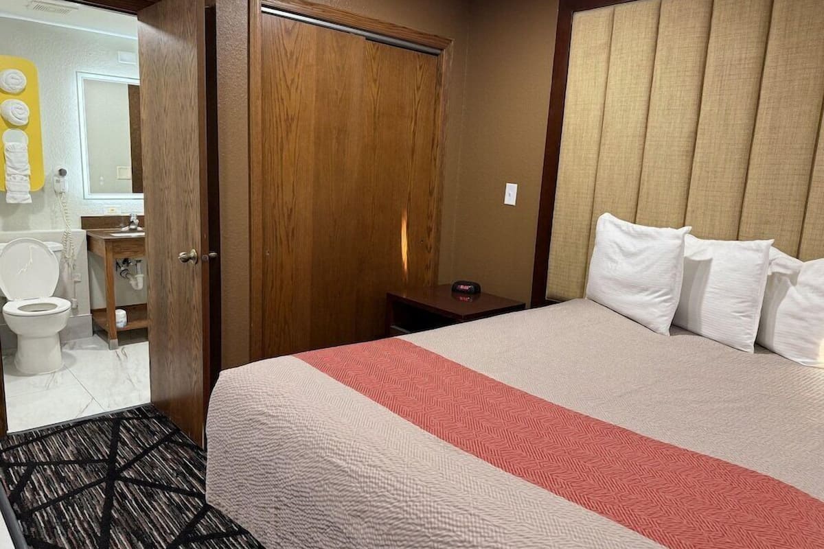 Una habitación de hotel con puerta de madera, cama con ropa de cama blanca y roja, mesita de noche y baño visible a través de una puerta abierta.