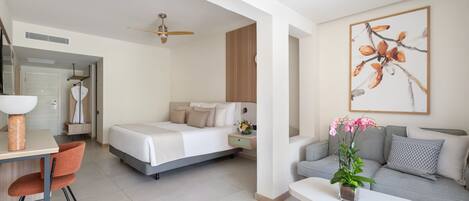 Une chambre d’hôtel moderne avec un lit, un canapé, un bureau et une chaise. On y trouve un ventilateur de plafond, un tableau au mur et une plante sur le bureau.