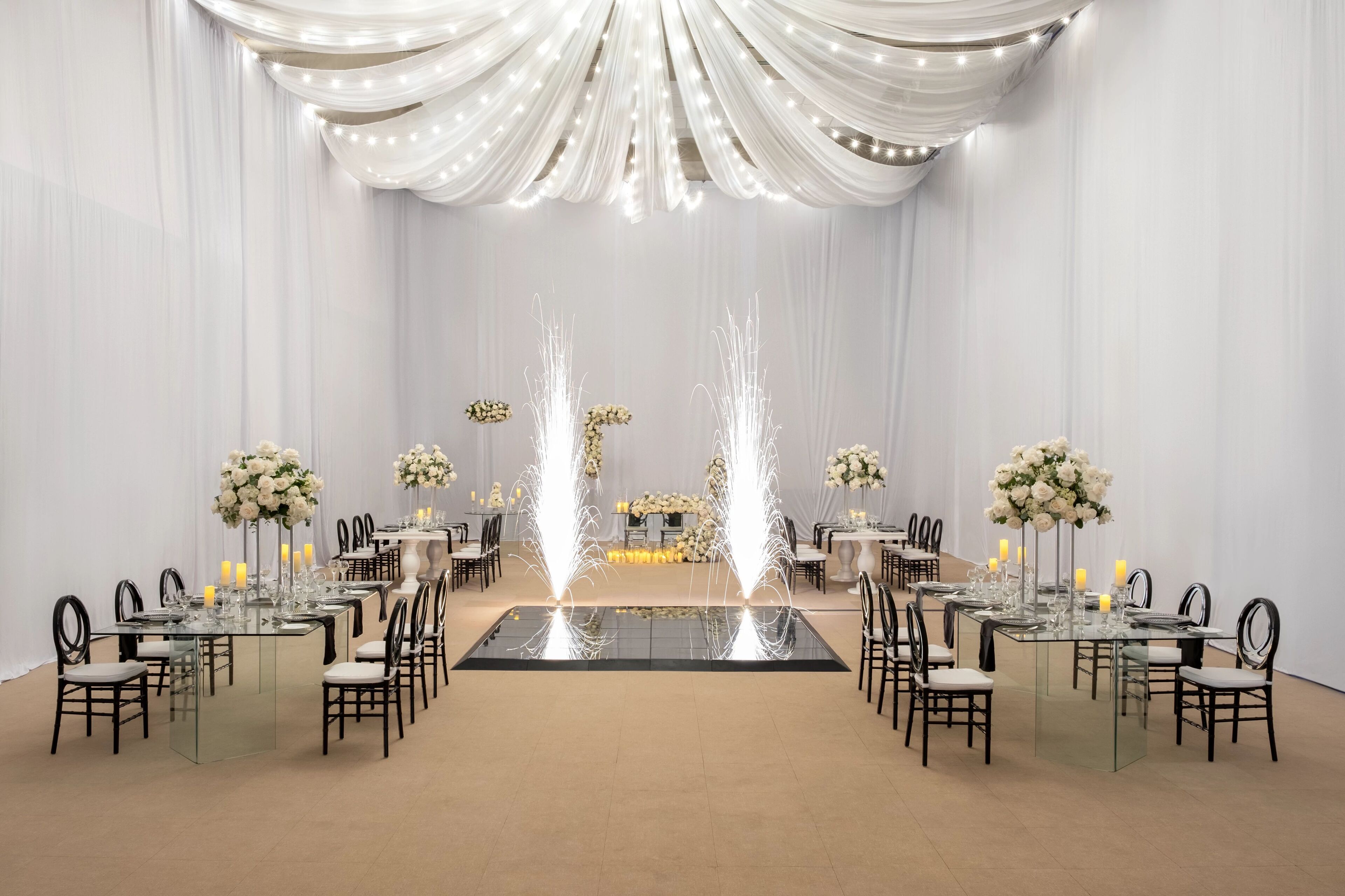 A formal event space with tables set for a banquet, featuring white drapery and floral centerpieces.