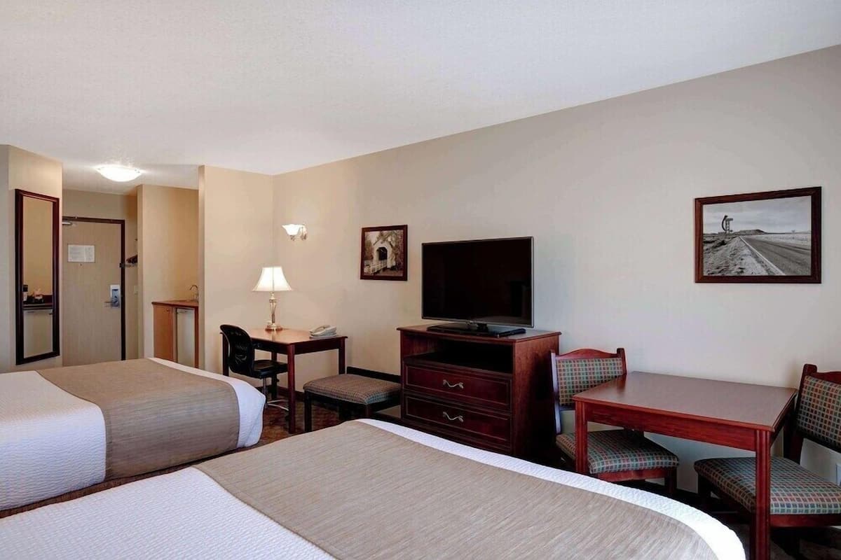 A hotel room with two beds, a desk with a lamp, a television, and a dining table with chairs.