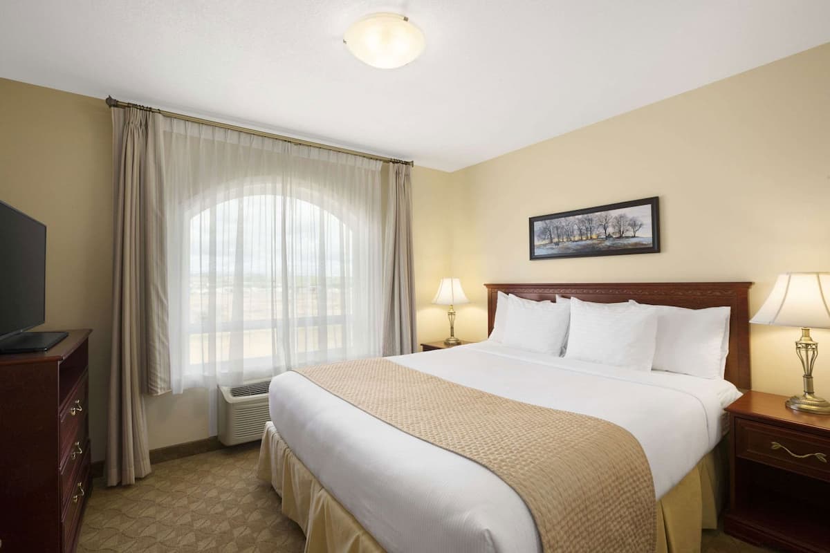 A hotel room with a large bed, a television, a desk, and a window with sheer curtains.
