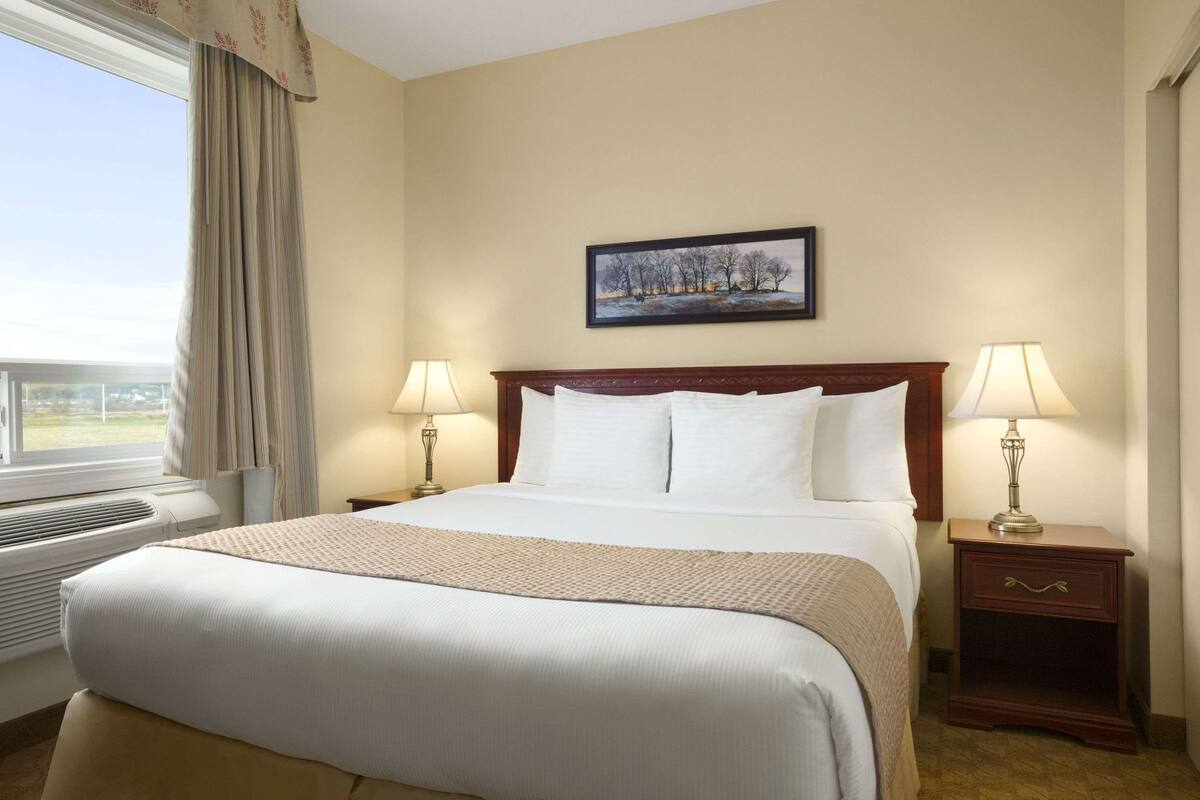 A hotel room with a large bed, two bedside tables with lamps, a window with curtains, and a painting on the wall.