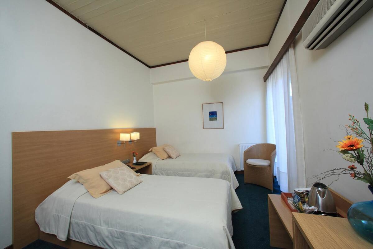 Habitación de hotel con dos camas, cabecera de madera, lámpara de pared, una silla de mimbre y un florero con flores.