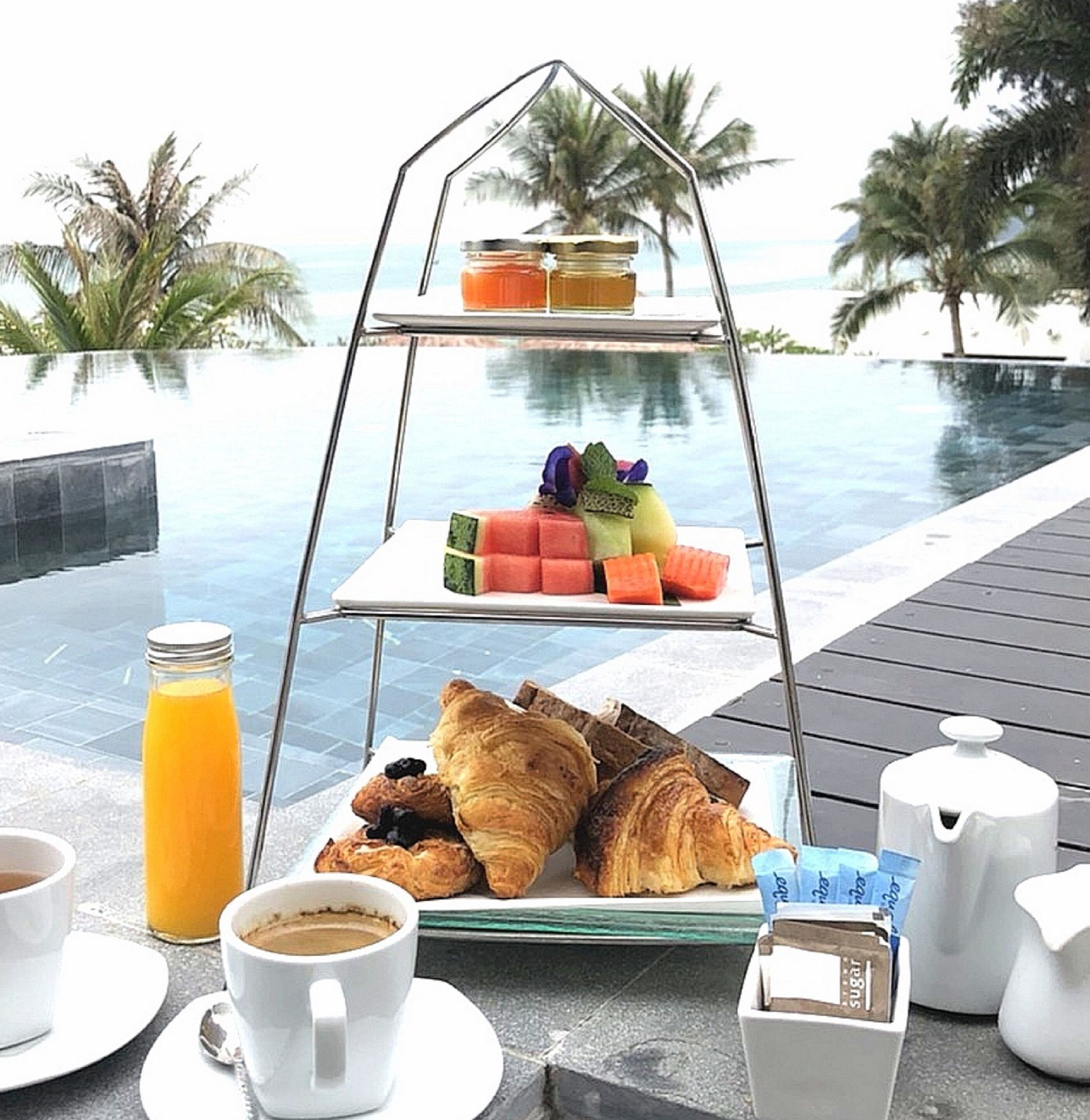A breakfast setup on a deck with a view of a pool and palm trees.
