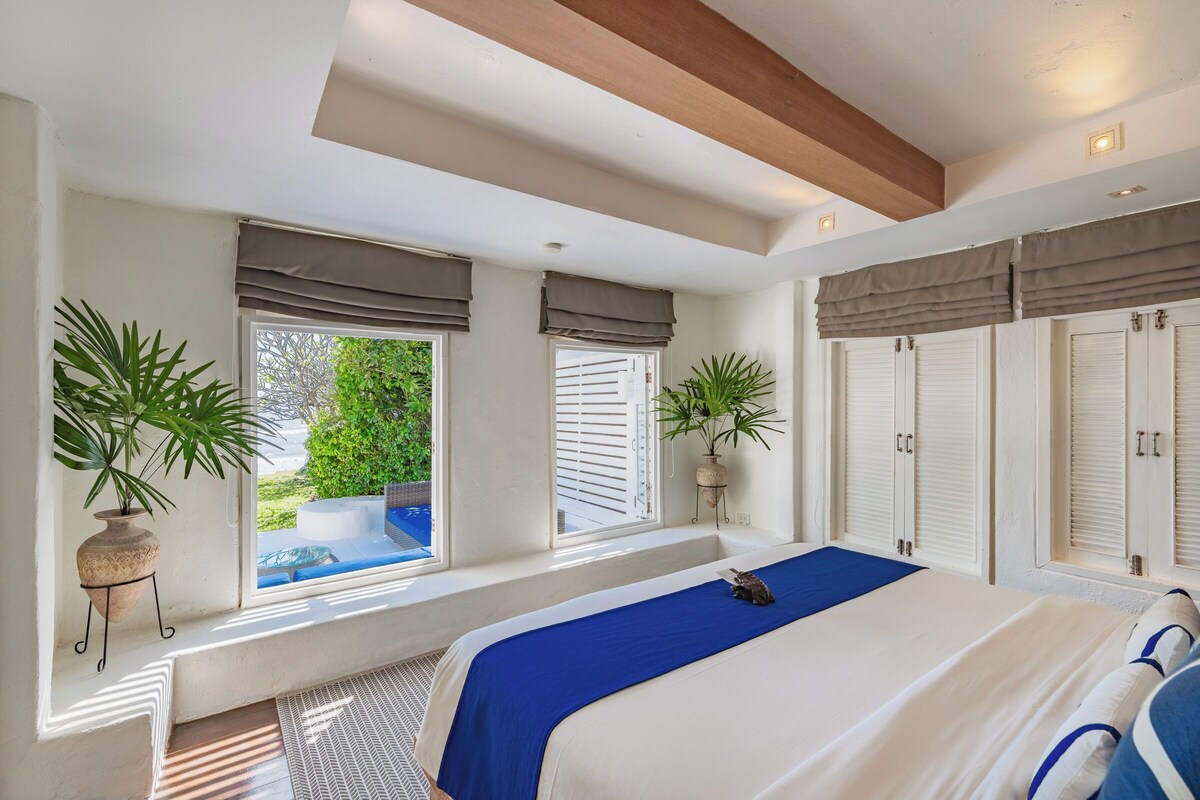 A bedroom with a large bed, a window with blinds, a potted plant, and a view of greenery outside.