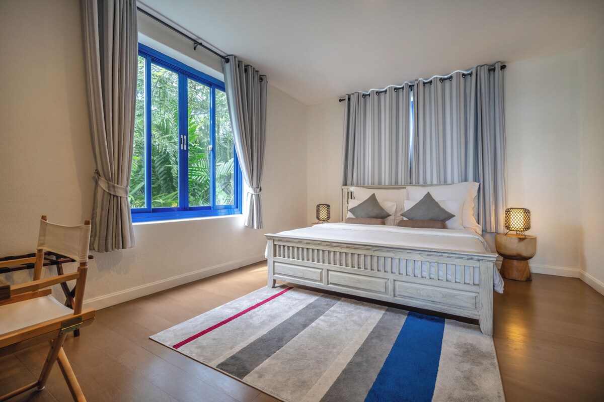 A bedroom with a large bed, a window with curtains, a nightstand with a lamp, and a striped rug.