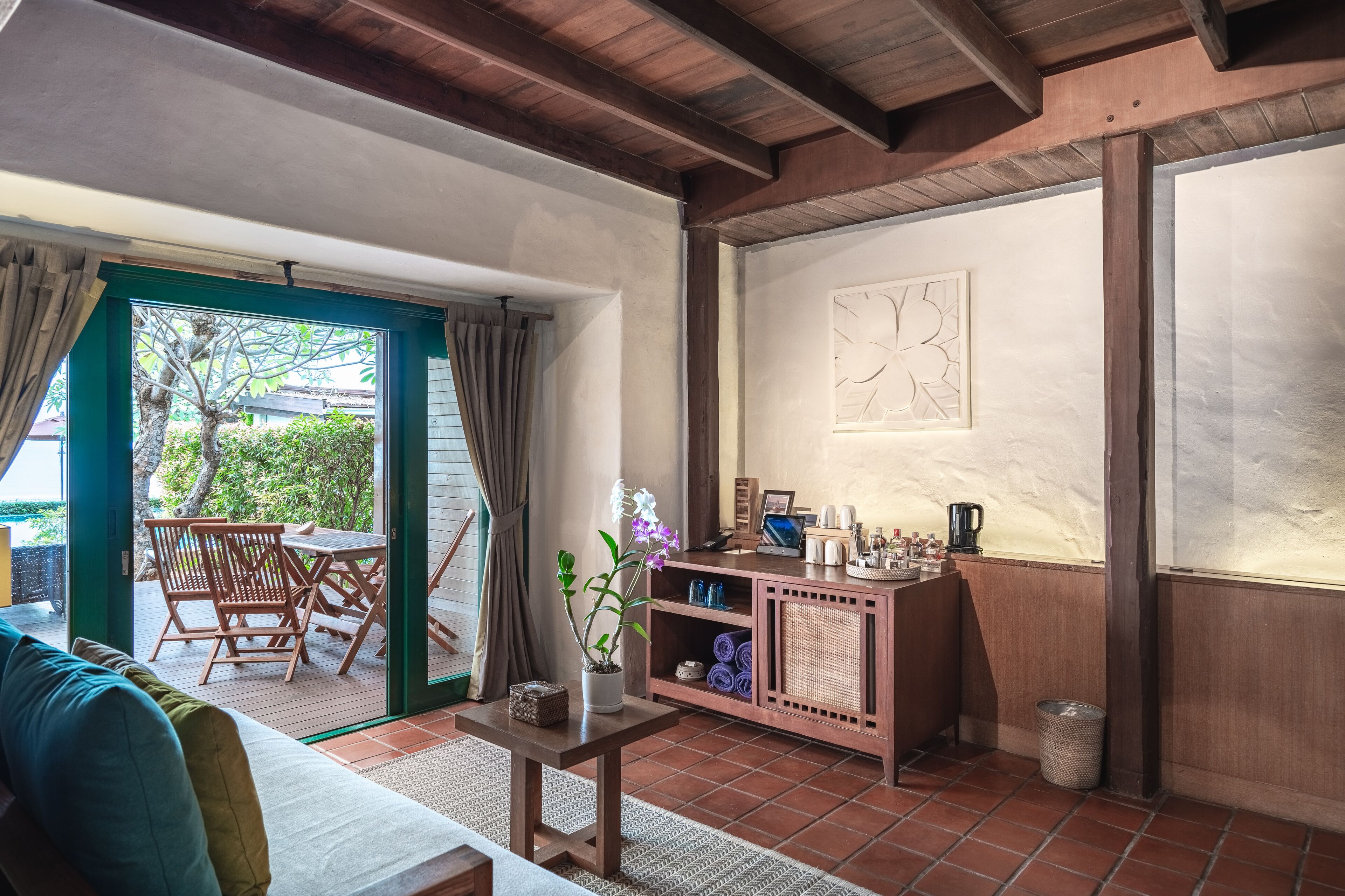 Una habitación acogedora con techo de madera, una zona de comedor con sillas y una mesa, un mueble pequeño con objetos, y vistas a través de un ventanal.