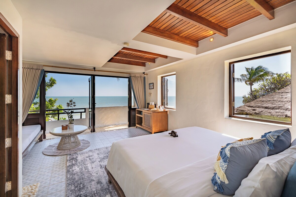 A bedroom with a large bed, a small table, a desk, and a view of the ocean.
