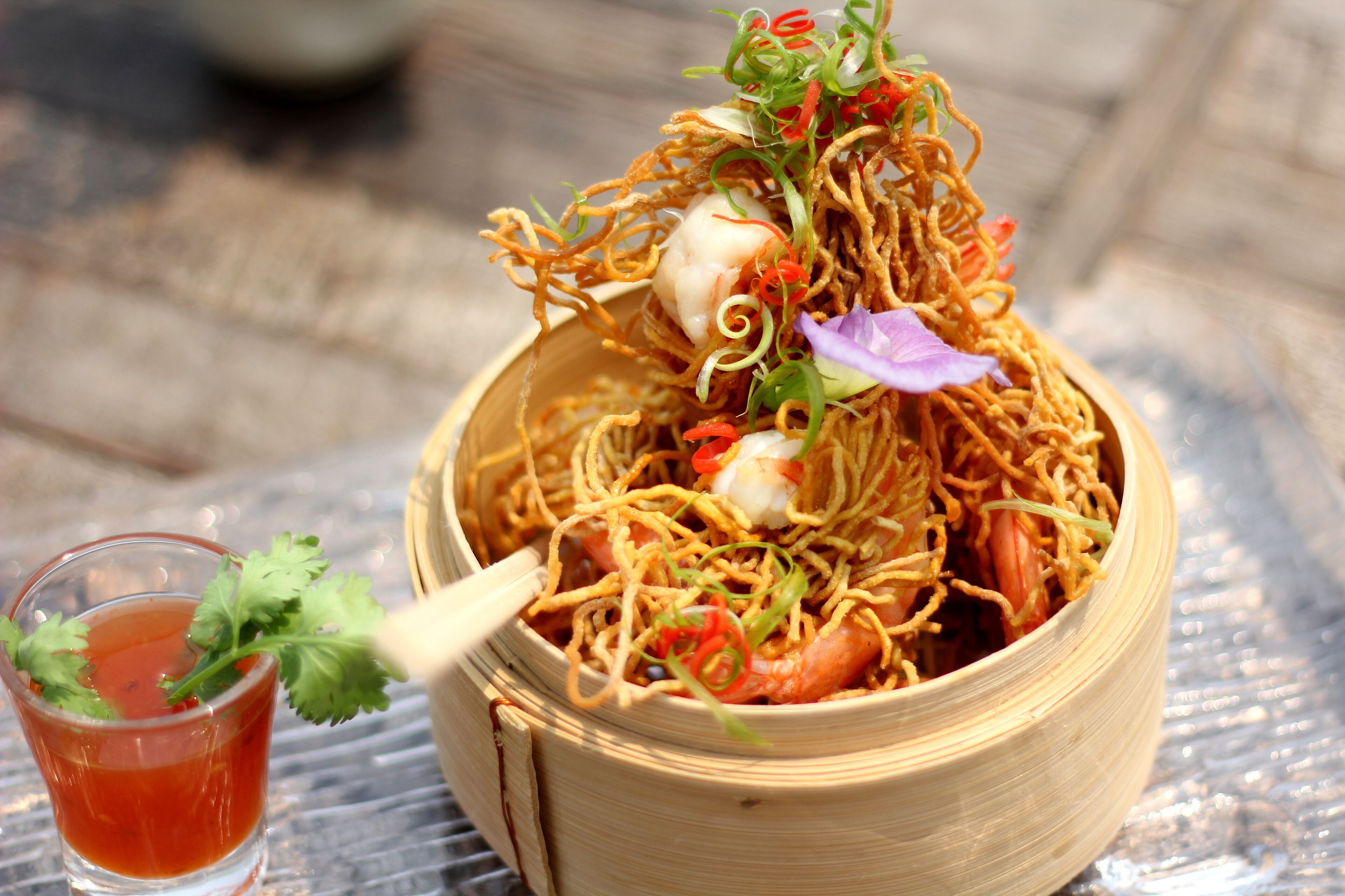 Een kom noodles met garnalen en groenten, een glas rode drank, en een lepel.